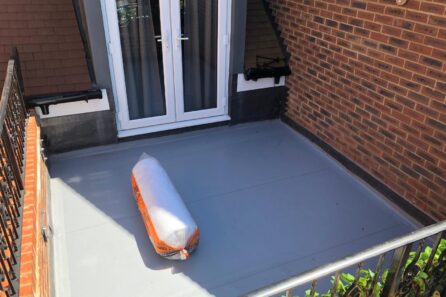 A grey flat roofing surface with a rolled-up roofing material positioned on it, located near a set of double doors.